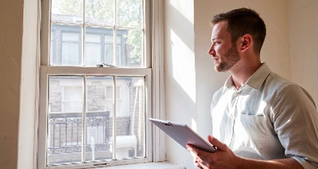 Energy assessor in a bright room holding a clipboard and looking out of a sash window